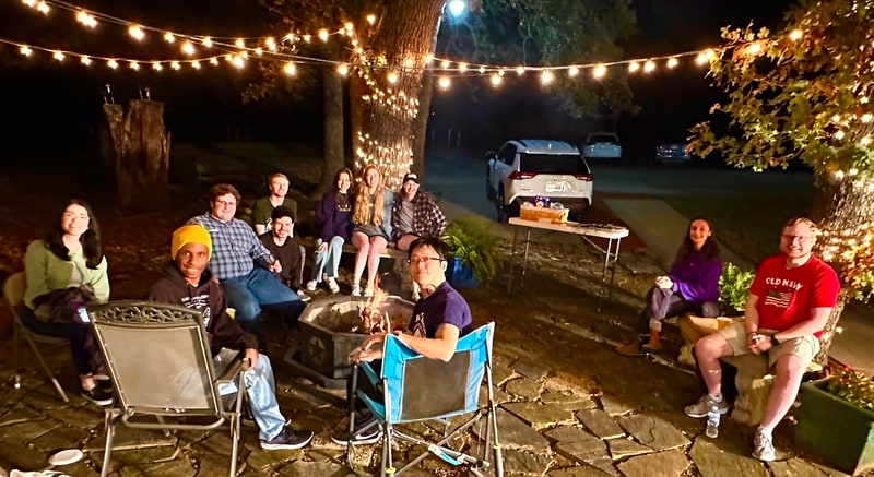 Friends sitting in a circle around a fire pit at night under strings of lights.