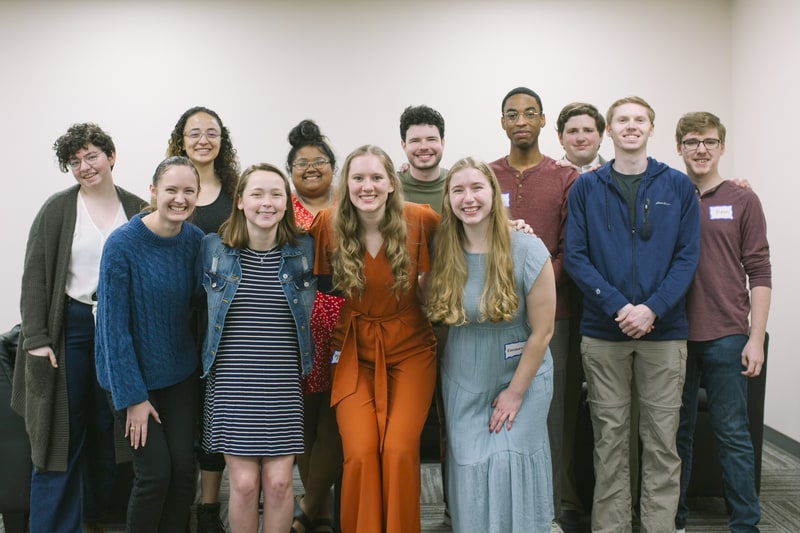 About a dozen young adults smiling together for a group photo against a plain wall.