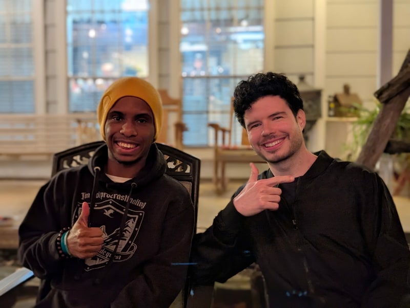 Two friends grinning on a porch in the evening, one giving a thumbs-up.