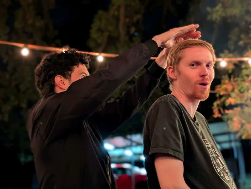 Two friends laughing outdoors at night under string lights, one holding a hand flat above the other’s head.