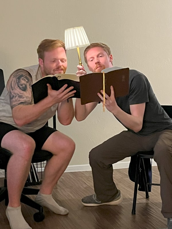 Two men leaning in over open Bibles, one holding up a lamp, hamming it up for the camera.